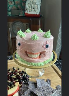 a pink cake with green icing and smiling face on a gold platter next to crackers