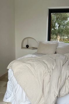 an unmade bed with white sheets and pillows in front of a large open window