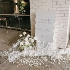 a white sign sitting on top of a floor next to a bunch of flowers in front of a brick wall