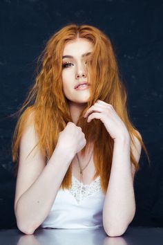 a woman with long red hair sitting at a table and posing for the camera in front of a black background