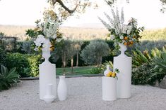 three white vases filled with flowers sitting on top of a gravel covered ground next to trees