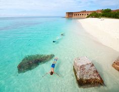 two people are swimming in the clear blue water near an old brick wall and beach