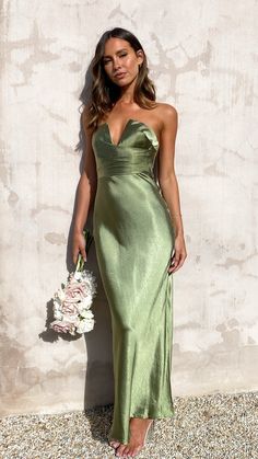 a woman in a green dress standing next to a white wall holding a flower bouquet