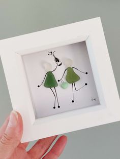 a hand holding up a small white frame with two paper cutouts of people in green dresses