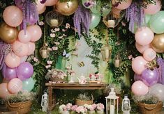 a table topped with lots of balloons next to a wall covered in flowers and greenery