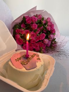 a birthday cake with a lit candle and pink roses in a paper box next to it