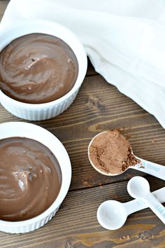 two white bowls filled with chocolate pudding next to spoons on a wooden table top