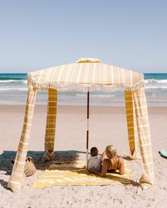 a woman and child sitting under an umbrella on the sand at the beach near the ocean