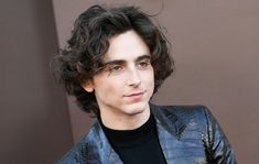 a young man with curly hair wearing a blue jacket and black turtle neck sweater looking at the camera