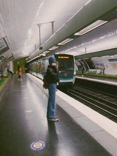 a person standing on the platform next to a train at a station with people walking by