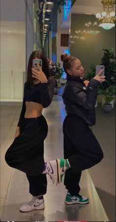 two young women taking pictures with their cell phones in an office building hallway, both wearing black clothing and green sneakers