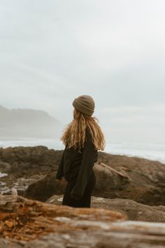 a woman standing on top of a rocky beach next to the ocean wearing a hat