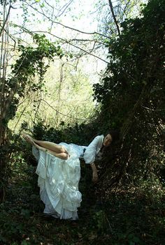 a woman in a white dress laying on the ground surrounded by trees and bushes with her arms stretched out