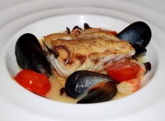 a white plate topped with fish and mussels covered in sauce on top of a table