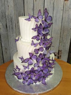 a three tiered white cake with purple butterflies on the top and bottom, sitting on a wooden table