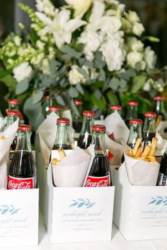 several sodas and french fries in small boxes on a table with flowers behind them