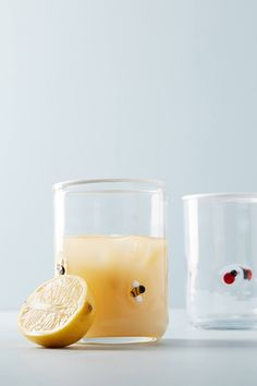 two glasses filled with liquid next to a sliced lemon on a white counter top, one has a piece of fruit in it and the other is half empty