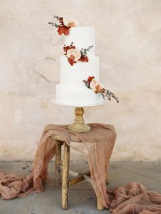 a white wedding cake with flowers on top sitting on a small stool next to a wall