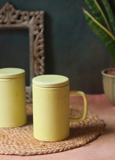 two yellow coffee mugs sitting on top of a woven place mat next to a potted plant