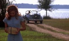 a woman holding a basket standing in front of a car on a dirt road next to a body of water