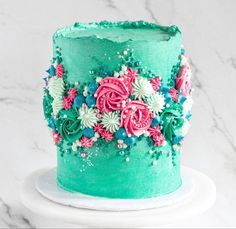 a green cake with pink and blue flowers on it sitting on a white pedestal in front of a marble background