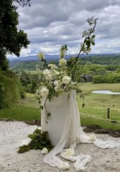 a large vase with flowers on top of a gravel ground next to a field and trees