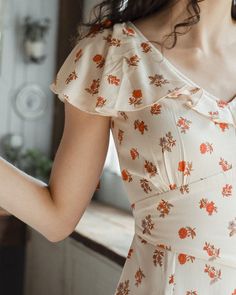 a woman in a floral print dress is standing near a window and looking at her cell phone