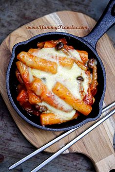 a skillet filled with pasta and sauce on top of a wooden cutting board next to utensils