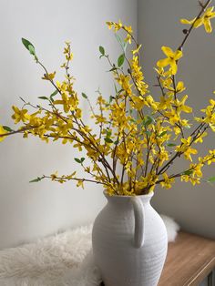 a white vase filled with yellow flowers on top of a wooden table next to a wall