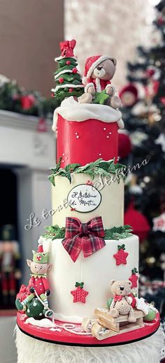 a three tiered christmas cake decorated with teddy bears and presents on top, sitting in front of a christmas tree