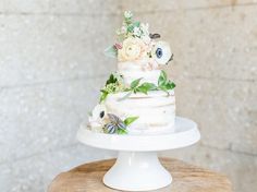 a three tiered cake with flowers on top sits on a wooden table next to a brick wall