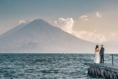 a bride and groom standing on a pier looking out at the ocean with a mountain in the background