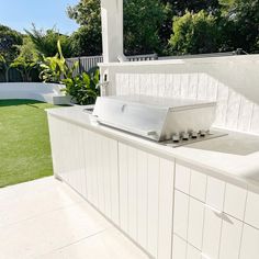 an outdoor kitchen with white tile counter tops and appliances on the outside wall, in front of a lush green yard