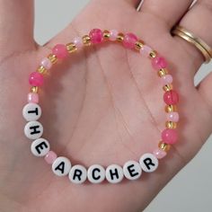 a hand holding a beaded bracelet with the word archer spelled in white and pink beads