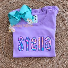 a purple shirt with the word stella on it and a blue bow is sitting on a woven mat