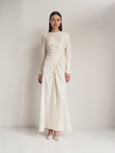 a woman standing in front of a white wall wearing a long sleeved dress with an asymmetrical design