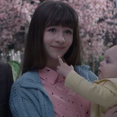 a woman holding a baby in her arms and looking at the camera with pink flowers behind her