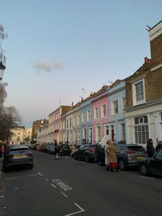 people are walking down the street in front of some colorful houses and parked cars on both sides