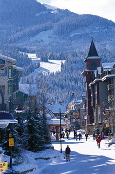 people are walking down the street in front of some buildings and snow covered mountains on a sunny day