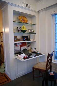 a desk and chair in a room with shelves on the wall above it, next to a window