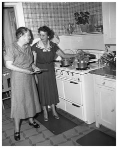 two women standing in a kitchen preparing food on top of stoves and pans