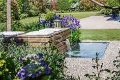 an outdoor hot tub surrounded by purple flowers and greenery in the foreground, next to a pool with water running through it