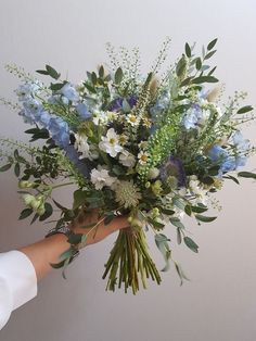 a person holding a bouquet of blue and white flowers with greenery in their hand