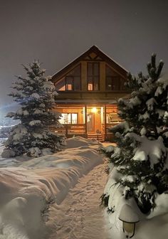 a cabin is lit up at night in the snow with pine trees on either side