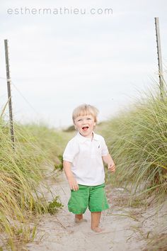 a little boy that is standing in the sand near some grass and bushes with his mouth open