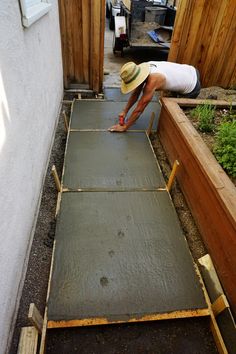 a man in a straw hat is working on a concrete slab near a fence and garden