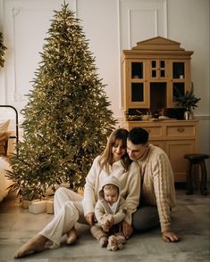 a man and woman are sitting next to a christmas tree with a baby on the floor