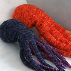 two knitted octopus toys sitting next to each other on a white counter top, one is orange and the other is purple