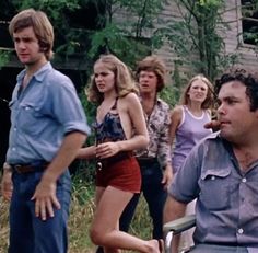 a group of people standing and sitting in front of an old house, one man is eating something