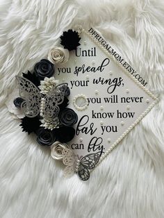 a graduation cap decorated with black and white paper flowers on a fluffy white blanket that says, until you spread your wings, you will never know how far you can fly